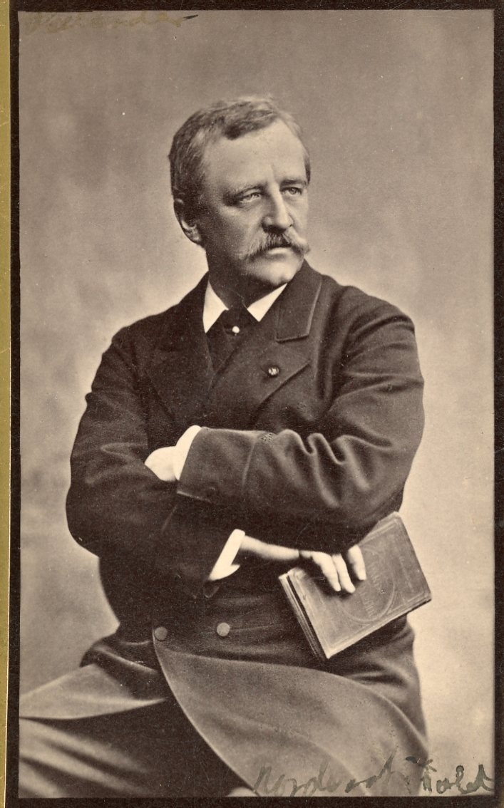 Kabinettsfotografi av en mustaschprydd man som sitter med armarna i kors. Han håller en bok i ena handen.