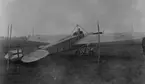 Flygplan M 2, Nieuport IV-G står på ett flygfält, inhängnad med rep. I förarsitsen sitter en man. Vy bakifrån.