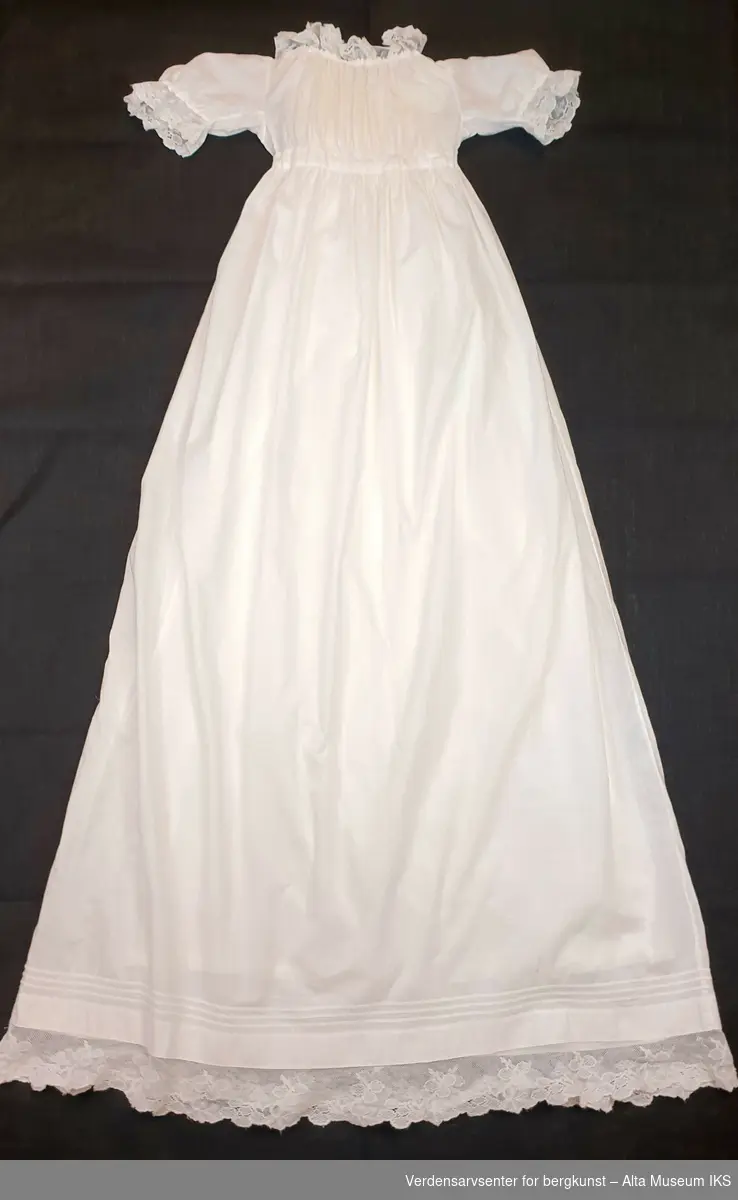 Hvit dåpskjole i bomull med blonder rundt hals, ermer og skjørtekant. Snøring med hvitt bånd i halslinning og rundt livet. Underkjole uten ermer i syntetisk materiale.