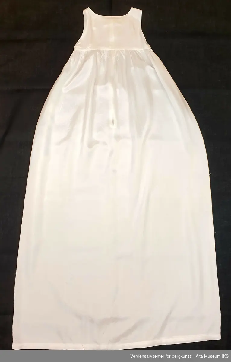 Hvit dåpskjole i bomull med blonder rundt hals, ermer og skjørtekant. Snøring med hvitt bånd i halslinning og rundt livet. Underkjole uten ermer i syntetisk materiale.