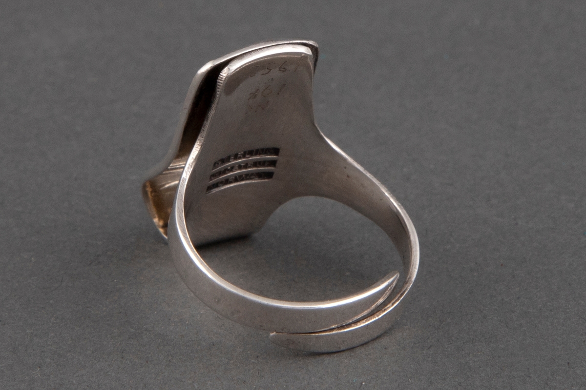 Industrielt fremstilt ring i sølv, utstanset i ett stykke og bøyet sammen. Ringplaten er av overbøyet rektangulær form og dekket av grønn halvgjennomsiktig emalje over et rutemønster med 8 utsparte rektangulære sølvpletter. Ringen er åpen på undersiden. De to ender krysser hverandre og gir utvidelsesmulighet.