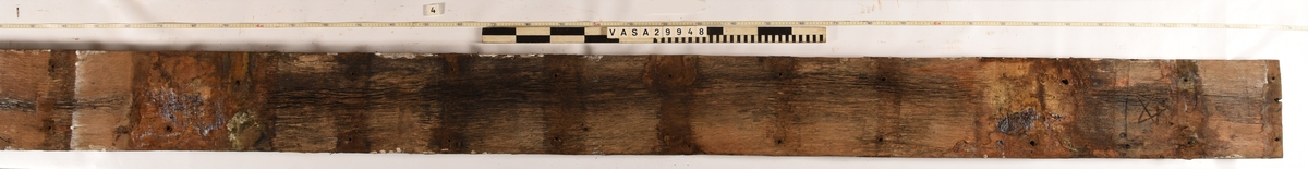 Däcksplanka i ek
Totalt 41genomgående spikhål, 20 längs plankans ena sida och 20 längs den andra. Ytterligare ett i mitten av ena änden där det också är en spricka.
Spikhålen är original från när skeppet byggdes. Originalspikarna är bortrostade. Efter bärgningen monterades däcksplankan med rostfri fransk träskruv samt bricka. Vid demonteringen 2022 lossades alla skruvar. De brickor som gick att ta bort togs bort. En del satt så djupt i träet att det inte gick att demontera dem utan att skada plankan. Dessa brickor fick därför sitta kvar.
Inristad stjärna i ena änden av plankans undersida.
Undersidan är täckt av små sprickor samt har tydliga avtryck från ribbor och däcksbalkar.