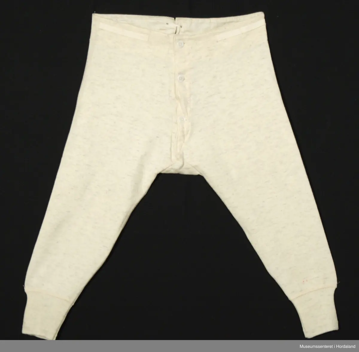 Lang ullunderbukse med åpning framme med band og tre knappar, og opning bak med justerbare bånd. Laga i "ren ull garantert", "norsk fabrikat".