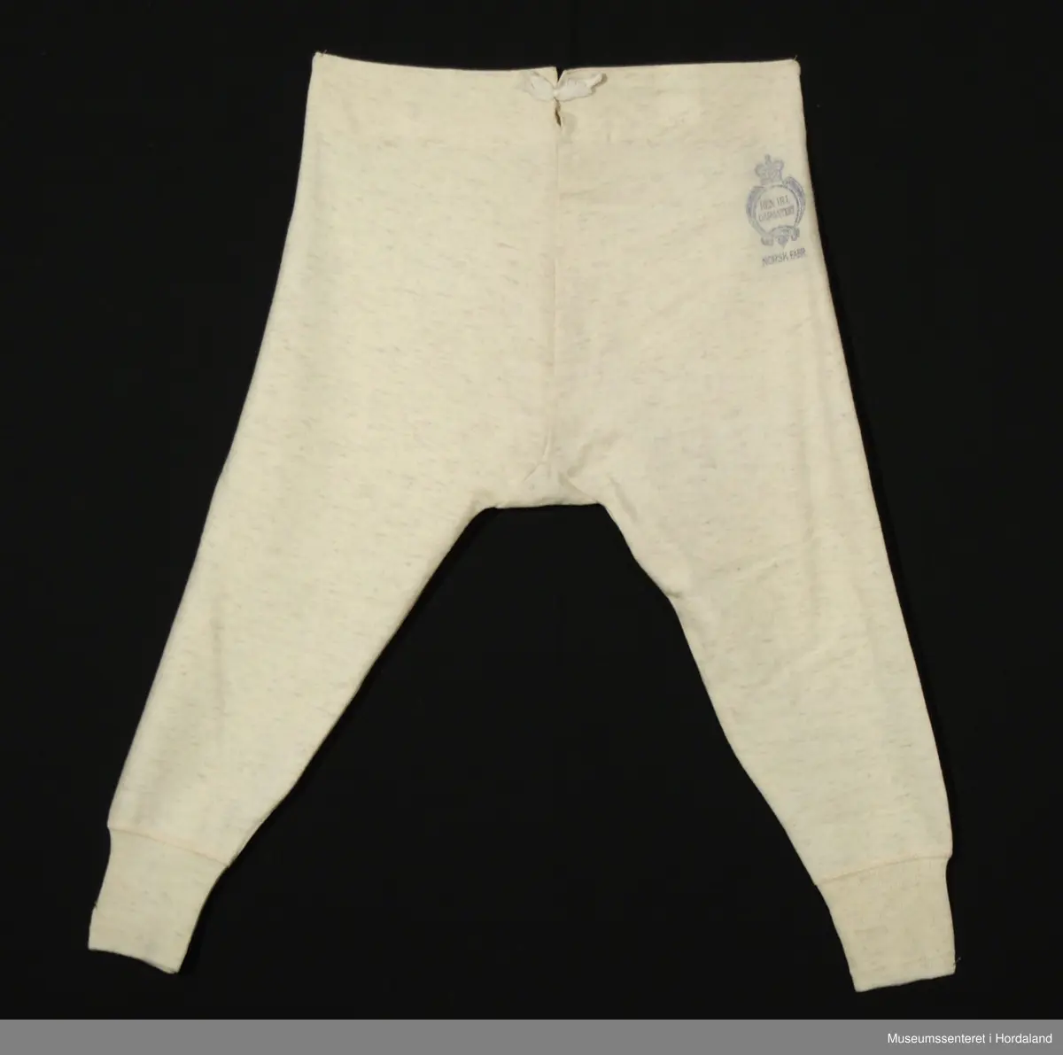 Lang ullunderbukse med åpning framme med band og tre knappar, og opning bak med justerbare bånd. Laga i "ren ull garantert", "norsk fabrikat".