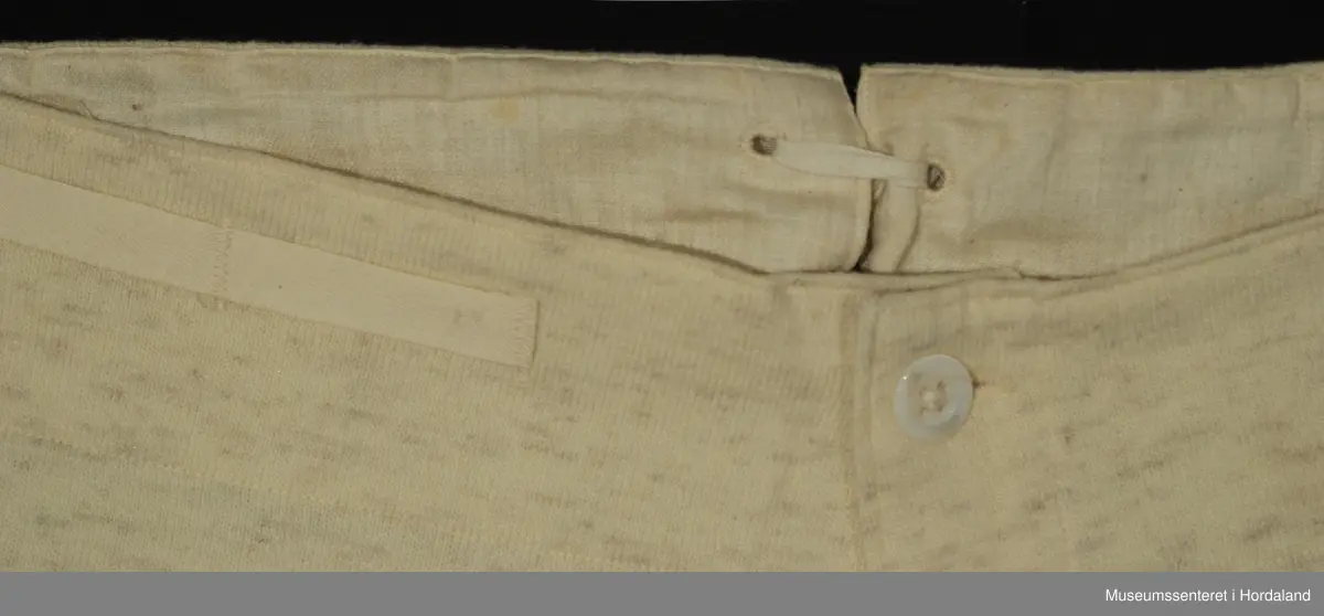 Lang ullunderbukse med åpning framme med band og tre knappar, og opning bak med justerbare bånd. Laga i "ren ull garantert", "norsk fabrikat".