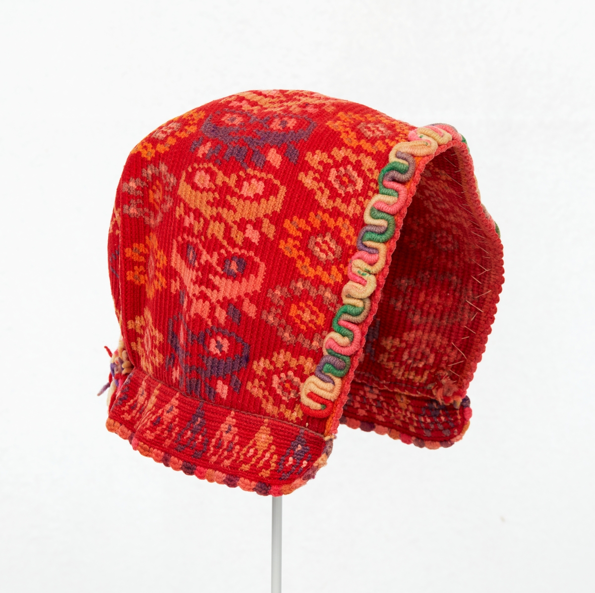 Hätta till Gagnefs kvinnliga sockendräkt. Flerfärgsvirkad av ullgarn i rött med mönster i lila, grått, gult och orange. En rund platta i nacken samt en påsydd 30 mm bred kant nertill. En smal röd uddspets virkad längst framkanten och en i lila, rosa och gult längst nederkanten. På framkanten påsytt en bågformad garnering av ullgarn i hättans färger. Mitt bak en tofs av samma garn.