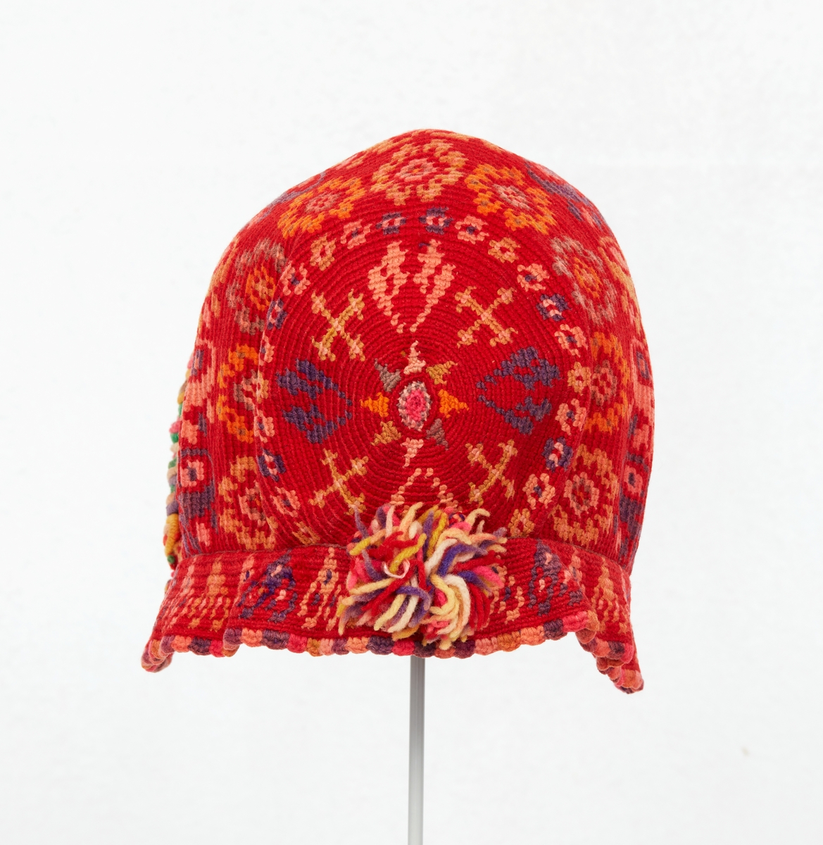 Hätta till Gagnefs kvinnliga sockendräkt. Flerfärgsvirkad av ullgarn i rött med mönster i lila, grått, gult och orange. En rund platta i nacken samt en påsydd 30 mm bred kant nertill. En smal röd uddspets virkad längst framkanten och en i lila, rosa och gult längst nederkanten. På framkanten påsytt en bågformad garnering av ullgarn i hättans färger. Mitt bak en tofs av samma garn.