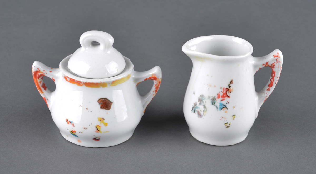 Sett med fløtemugge og sukkerskål.
Muggen har hanke med delvis avslitt rød dekor og et dekorbilde på ene siden, som er delvis slitt av. 
Sukkerskåla har håndtak på hver side og et lokk. Rød dekorlinje rundt åpningen på på hankene, som er delvis slitt av. 
Sukkerskåla har også dekorbilde på siden, som er nesten slitt av.