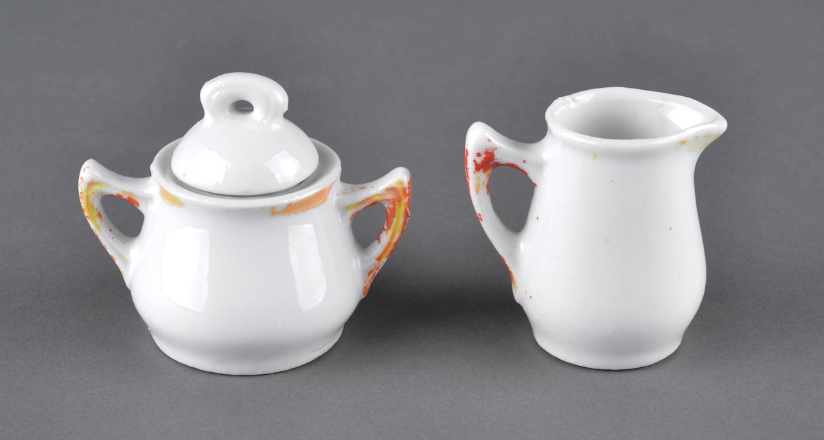 Sett med fløtemugge og sukkerskål.
Muggen har hanke med delvis avslitt rød dekor og et dekorbilde på ene siden, som er delvis slitt av. 
Sukkerskåla har håndtak på hver side og et lokk. Rød dekorlinje rundt åpningen på på hankene, som er delvis slitt av. 
Sukkerskåla har også dekorbilde på siden, som er nesten slitt av.