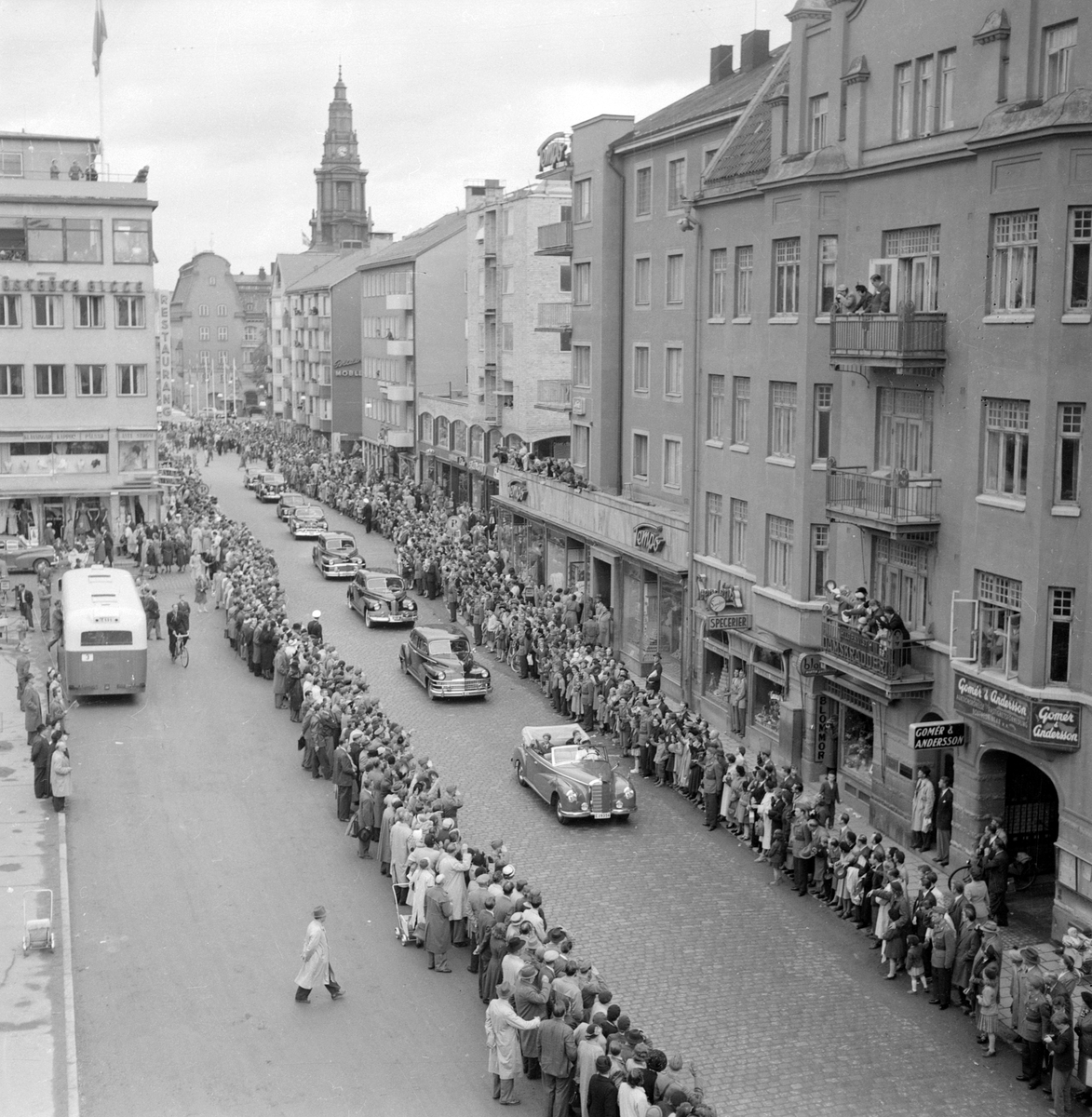 Kunglig kortege på Sankt Larsgatan. Kung Gustav VI Adolf och drottning Louise åker i samband med sin Eriksgata i maj 1953 i en öppen Mercedes genom stadskärnan. Längs med gatan trängs åskådare. Linköpingsbesöket var mycket uppskattat och tidningsrubrikerna talade om "Louise-feber".