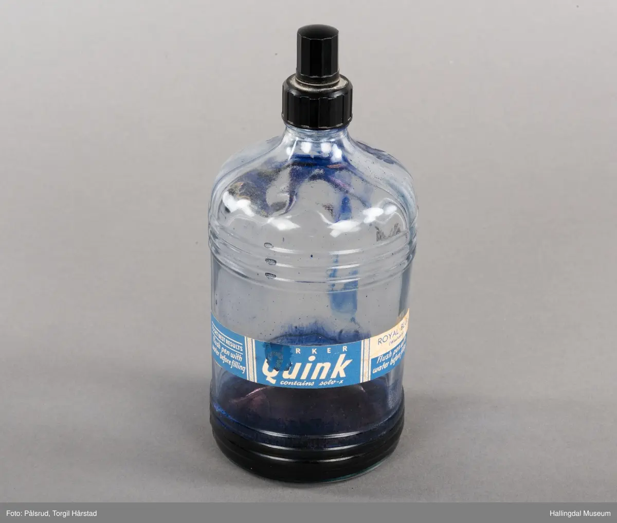 Flaske av blankt glas. Rett, sirkulært tverrsnitt. Det er litt blekk på flaska. Skruvekork av svart plast.