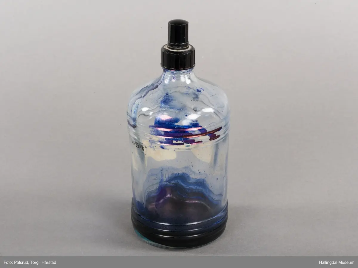 Flaske av blankt glas. Rett, sirkulært tverrsnitt. Det er litt blekk på flaska. Skruvekork av svart plast.