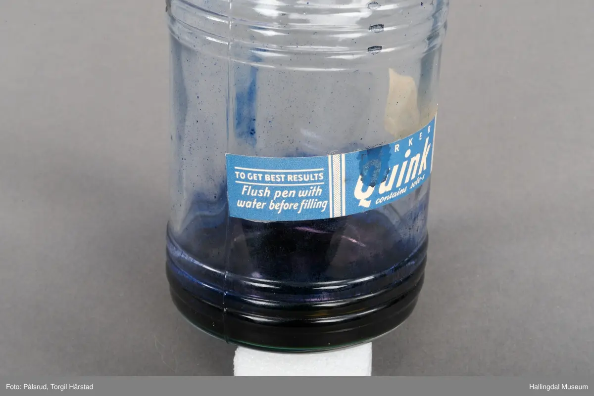 Flaske av blankt glas. Rett, sirkulært tverrsnitt. Det er litt blekk på flaska. Skruvekork av svart plast.