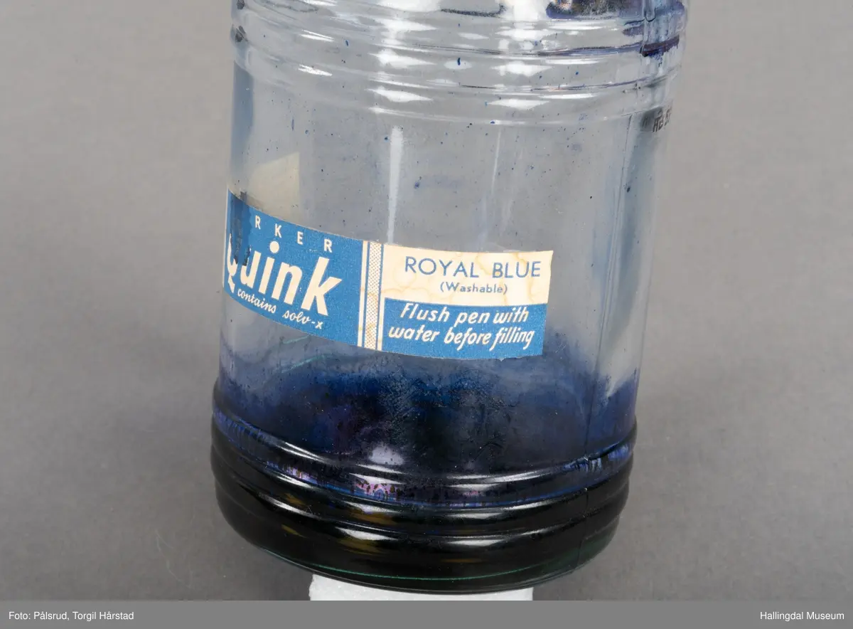 Flaske av blankt glas. Rett, sirkulært tverrsnitt. Det er litt blekk på flaska. Skruvekork av svart plast.