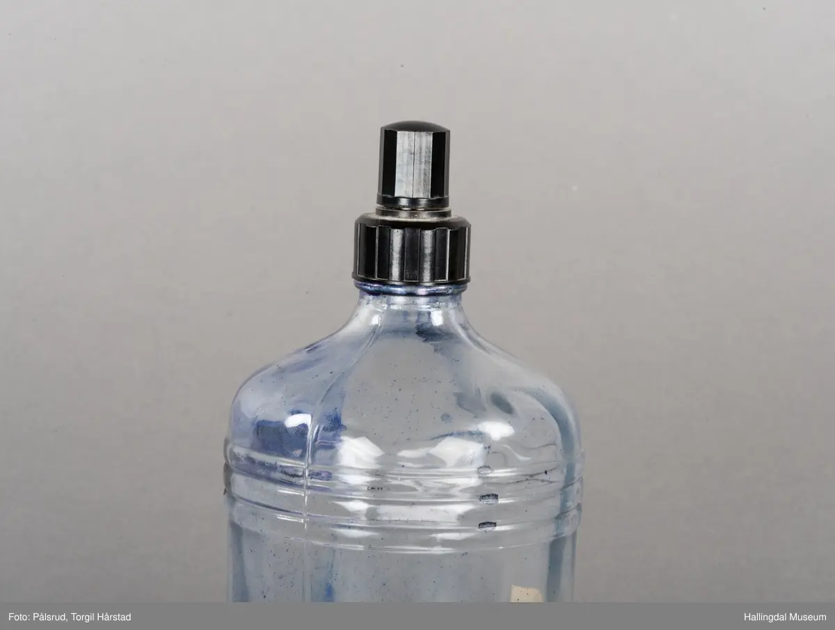 Flaske av blankt glas. Rett, sirkulært tverrsnitt. Det er litt blekk på flaska. Skruvekork av svart plast.