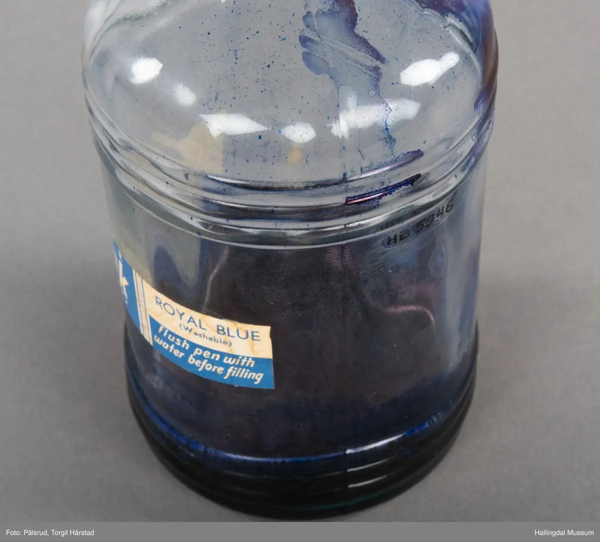 Flaske av blankt glas. Rett, sirkulært tverrsnitt. Det er litt blekk på flaska. Skruvekork av svart plast.