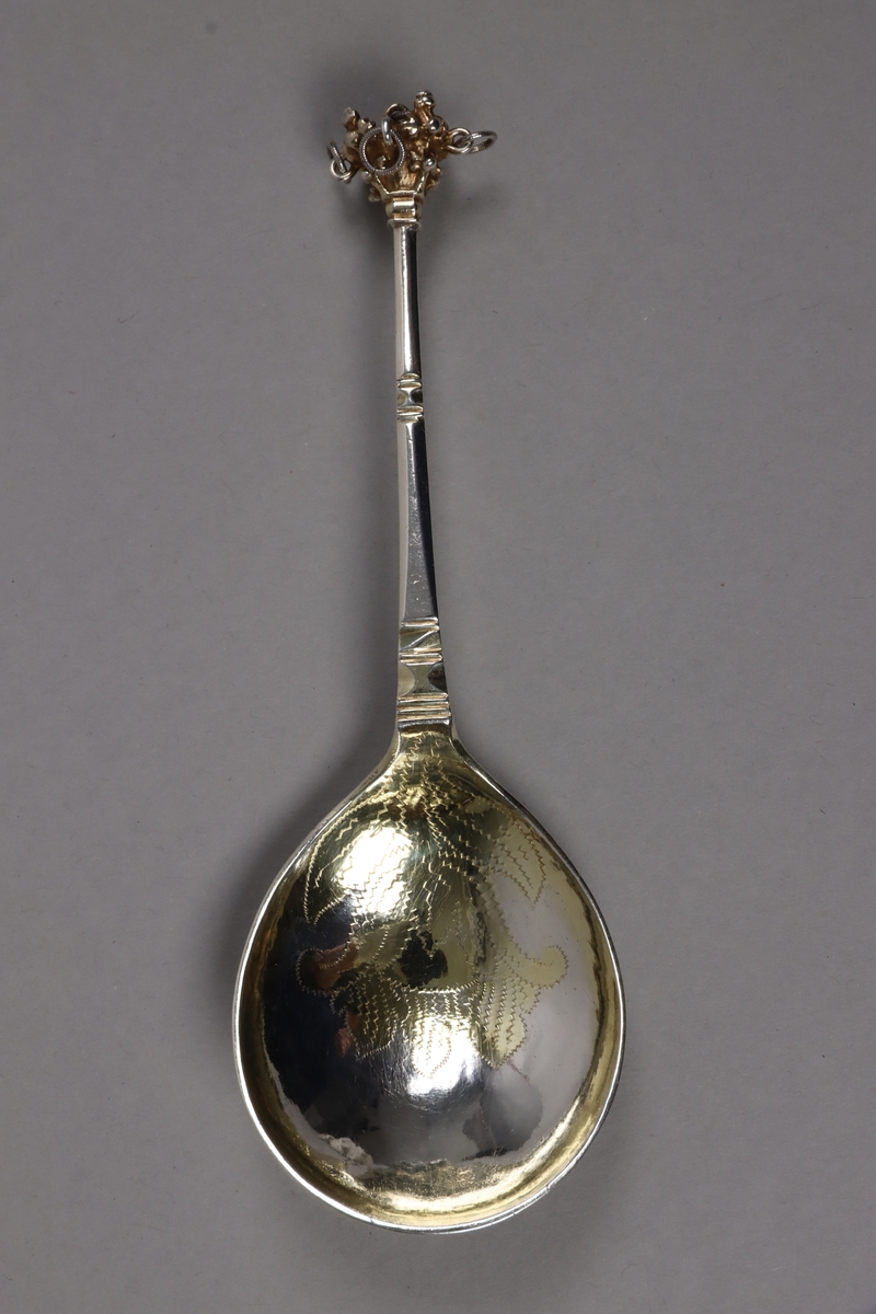 Brännvinssked av silver, med förgyllt skedblad och förgyllt dekorelement på handtaget. Skeden är utsågad ur plåt och har sedan fått drivet och graverat skedblad samt slagen ytdekor på handtaget. I änden sitter ett gjutet dekorelement med fem fastlödda ringar, i vilka sitter varsin större silverring och skramlar lite. Graverat på skedbladets undersida: NNS BED (delvis svårtytt). Stämplat på handtagets undersida: (svårtytt) stadsstämpel -  bockhuvud, CBM, A.