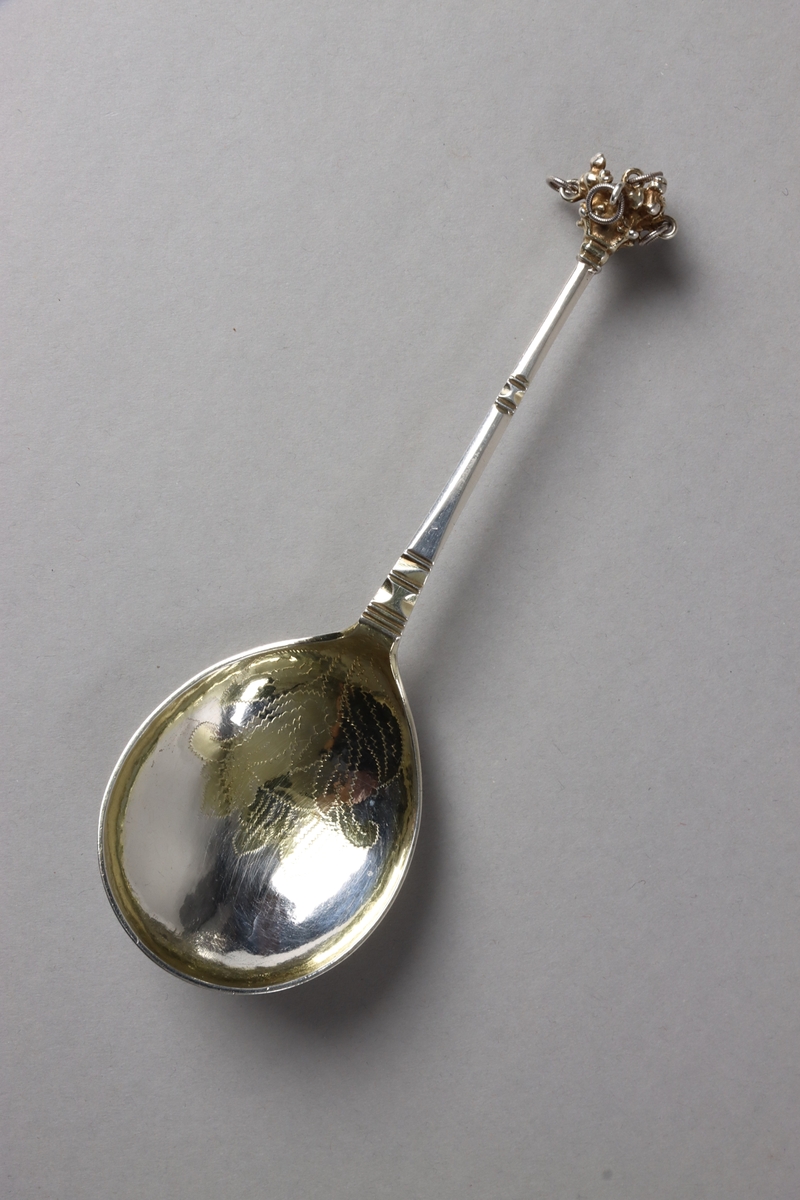 Brännvinssked av silver, med förgyllt skedblad och förgyllt dekorelement på handtaget. Skeden är utsågad ur plåt och har sedan fått drivet och graverat skedblad samt slagen ytdekor på handtaget. I änden sitter ett gjutet dekorelement med fem fastlödda ringar, i vilka sitter varsin större silverring och skramlar lite. Graverat på skedbladets undersida: NNS BED (delvis svårtytt). Stämplat på handtagets undersida: (svårtytt) stadsstämpel -  bockhuvud, CBM, A.