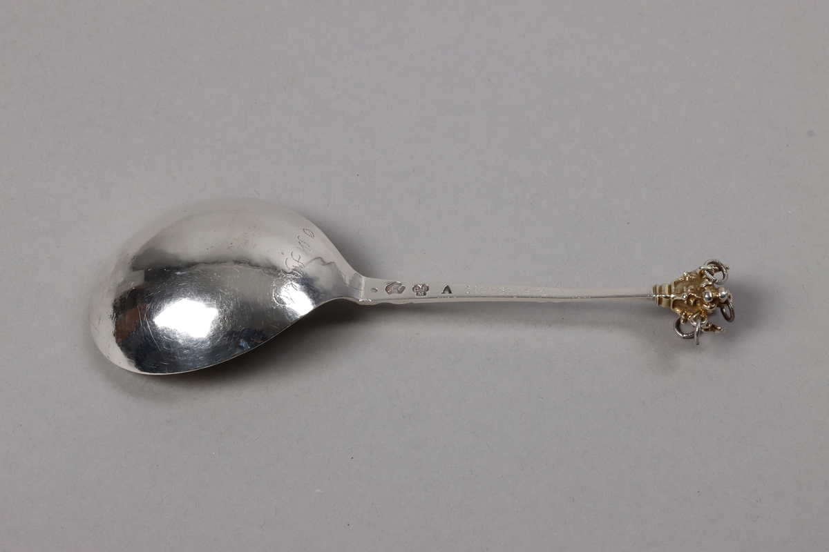 Brännvinssked av silver, med förgyllt skedblad och förgyllt dekorelement på handtaget. Skeden är utsågad ur plåt och har sedan fått drivet och graverat skedblad samt slagen ytdekor på handtaget. I änden sitter ett gjutet dekorelement med fem fastlödda ringar, i vilka sitter varsin större silverring och skramlar lite. Graverat på skedbladets undersida: NNS BED (delvis svårtytt). Stämplat på handtagets undersida: (svårtytt) stadsstämpel -  bockhuvud, CBM, A.