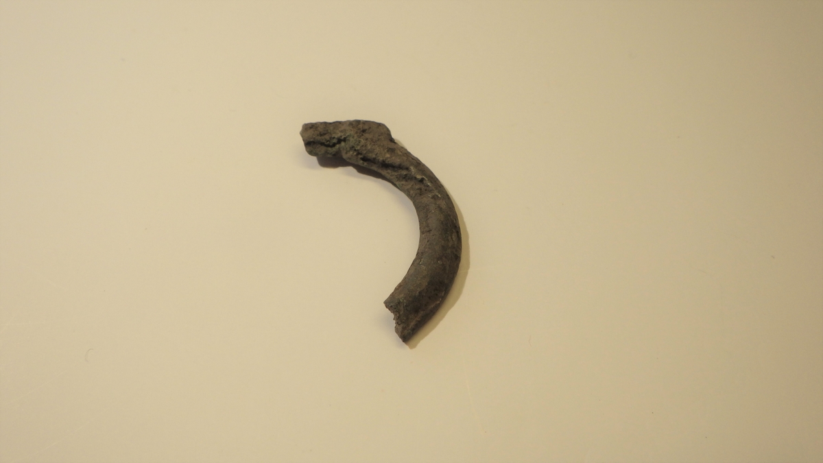 Del av beltespenne. Spennen er brukket og er tydelig mislykket i støpingen, med fordypninger og sprekker i godset. Tilnærmet flat underside og hvelvet overside. Som Geoff Eagan: Dress Accessories 1150-1450, tilnærmet fig. 279, 282 men uten påvislig leppe.