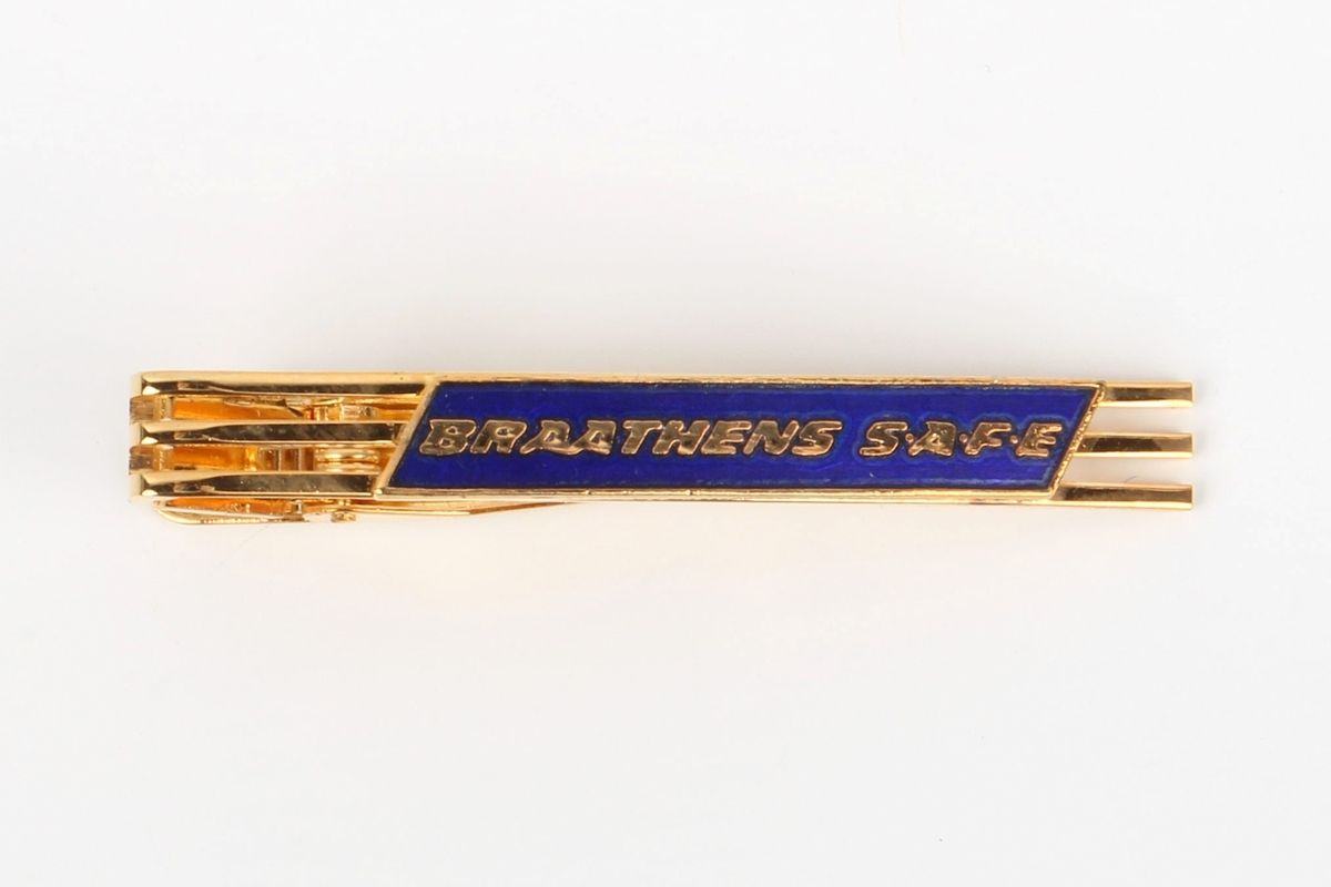 Gullfarget slipsnål med Braathens-logo i blå emalje. Festet på et stykke blå kartong, ligger i lite etui av plast med svart bunn og gjennomsiktig lokk.