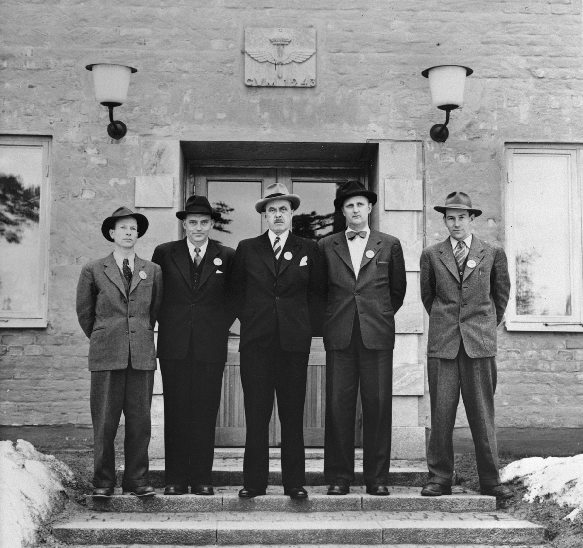 Gruppfoto av företagsledningen vid instrumentverksaden på Centrala verkstäderna Malmslätt, 1951. Fem män uppställda på kanslihusets trappa. Från vänster chef för kontrollavdelningen R.A. Sandberg, laboratoriechef Artur Fransson, styresman Harald Larsson, överingenjör Sture Edvardsson samt motoringenjör Gunnar Wikner från motoravdelningen.