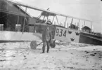 En militär flygare står vid flygplan Albatros SW 20 nummer 934, framför B-hangaren på Flygkompaniets tygverkstäder på Malmen. Vintertid, omkring 1919-1921. Motorprovbockarna till höger.