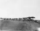 Sex flygplan Phönix D. II och en FVM Tummeliten uppställda på flygfältet vid Flygkompaniets tygverkstäder på Malmen, omkring 1920-1924. A- och B-hangarerna i bakgrunden.