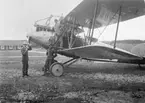 Två civilklädda och en militär poserar vid flygplan FVM S 18 på Flygkompaniets tygverkstäder på Malmen, omkring 1920. I bakgrunden syns filarverkstaden.