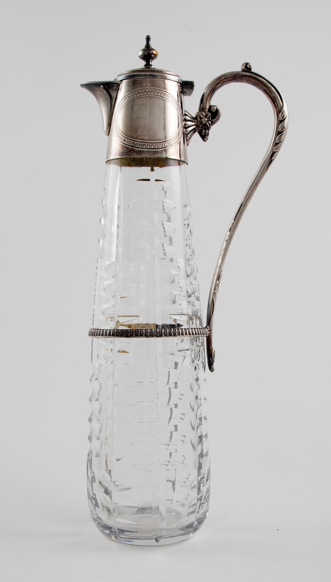 Høyreist kanne i klart glass med påmontert hank og lokk i sølvplett. Konisk korpus som er dekorert med sliping, i form av vertikale linjer som flankeres av parvis olivensliping. Svungen hank i sølvplett med blomsterdekor. Plettmonteringen på kannens øvre del har graverte ovaler, samt innpunslet inskripsjon: "Erindring / af / 23.9.82". Mykt buet nebb samt henglset lokk med balusterformet lokknapp.