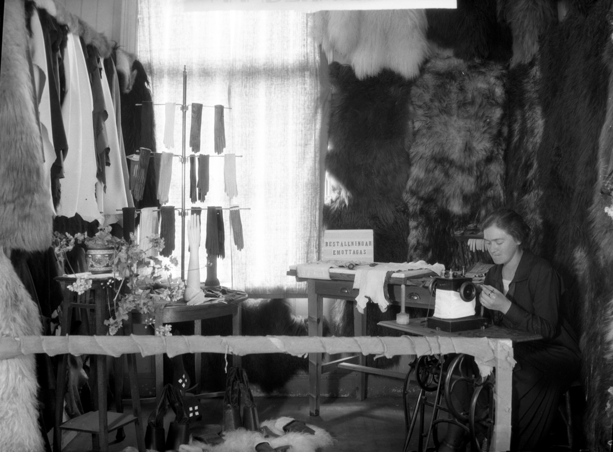 En sömmerska vid symaskinen i Ekengrens Handskmakeris utställningsmonter på Hantverksutställningen i Gamlebyskolan, Varberg 1927. I montern hänger pälsar, skinn och en ställning med handskar. Skylten förkunnar "Beställningar emottagas".