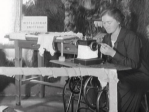 En sömmerska vid symaskinen i Ekengrens Handskmakeris utställningsmonter på Hantverksutställningen i Gamlebyskolan, Varberg 1927. I montern hänger pälsar, skinn och en ställning med handskar. Skylten förkunnar "Beställningar emottagas".