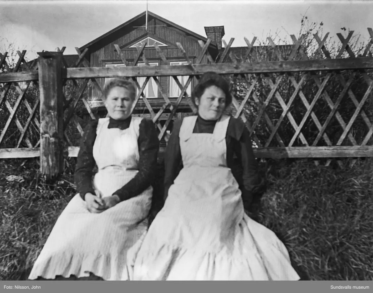Två pigor hos familjen Sahlén på Roken 1:3, pigan till höger är Margareta Johanna Sjöström. Gårdens mangårdsbyggnad syns bakom staketet.