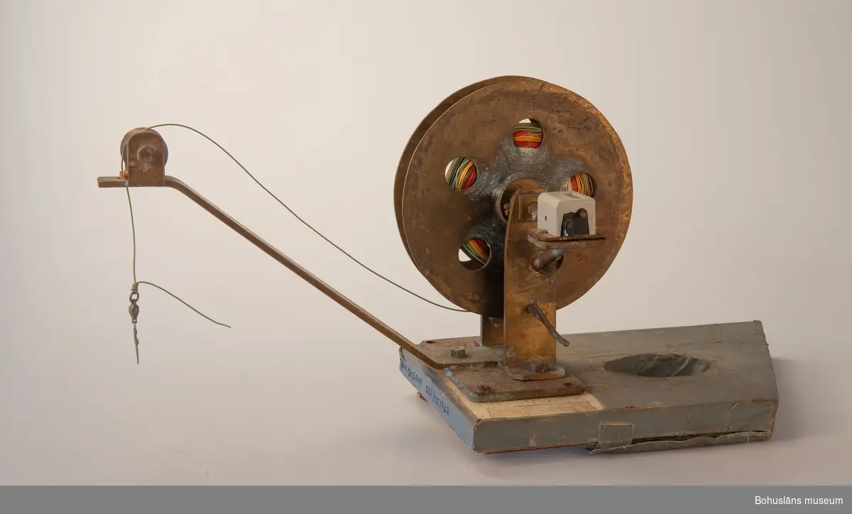 Hemmagjor manuell djuprigg. Fast bom av mässing och rulle av metall och vev med korkhandtag fästad mot en gråmålad bottenplatta. 
På rullen sitter flerfärgad nylonlina. I bottenplatta urtag för spö. Tejpad med grå tejp.
Räkneverk för  - troligen - antalet vevtag.