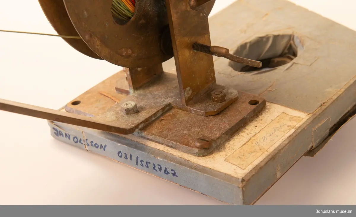 Hemmagjor manuell djuprigg. Fast bom av mässing och rulle av metall och vev med korkhandtag fästad mot en gråmålad bottenplatta. 
På rullen sitter flerfärgad nylonlina. I bottenplatta urtag för spö. Tejpad med grå tejp.
Räkneverk för  - troligen - antalet vevtag.