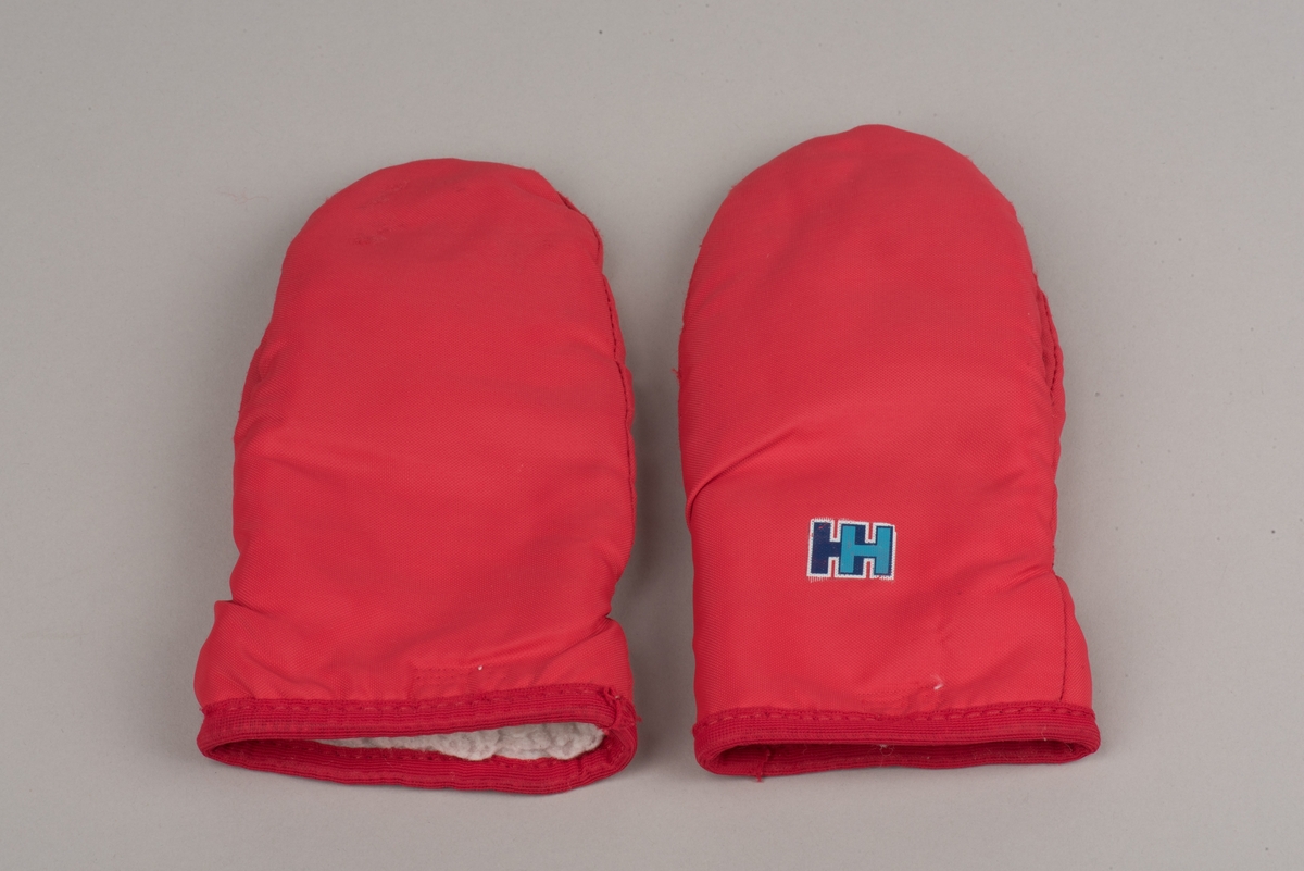 Röda tumvantar av polyester för barn av märket Helly Hansen.
Yttertyg av röd polyester och inuti ett foder av vitt fleecetyg.
På vänster vantes utsida finns Helly Hansens märke "HH" i två blåa nyanser med vit kant.
Vantarna är välanvända och jämnt slitna. De etiketter som finns inuti vantarna är blekta och svårlästa.