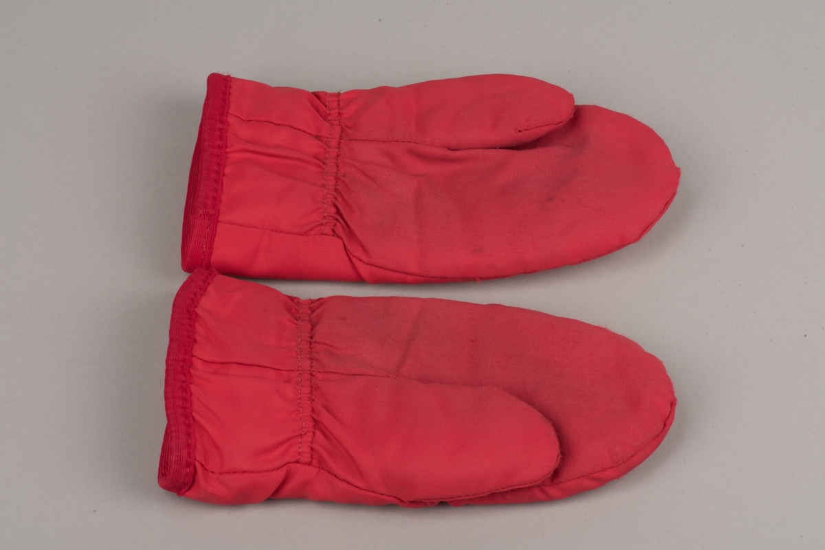 Röda tumvantar av polyester för barn av märket Helly Hansen.
Yttertyg av röd polyester och inuti ett foder av vitt fleecetyg.
På vänster vantes utsida finns Helly Hansens märke "HH" i två blåa nyanser med vit kant.
Vantarna är välanvända och jämnt slitna. De etiketter som finns inuti vantarna är blekta och svårlästa.