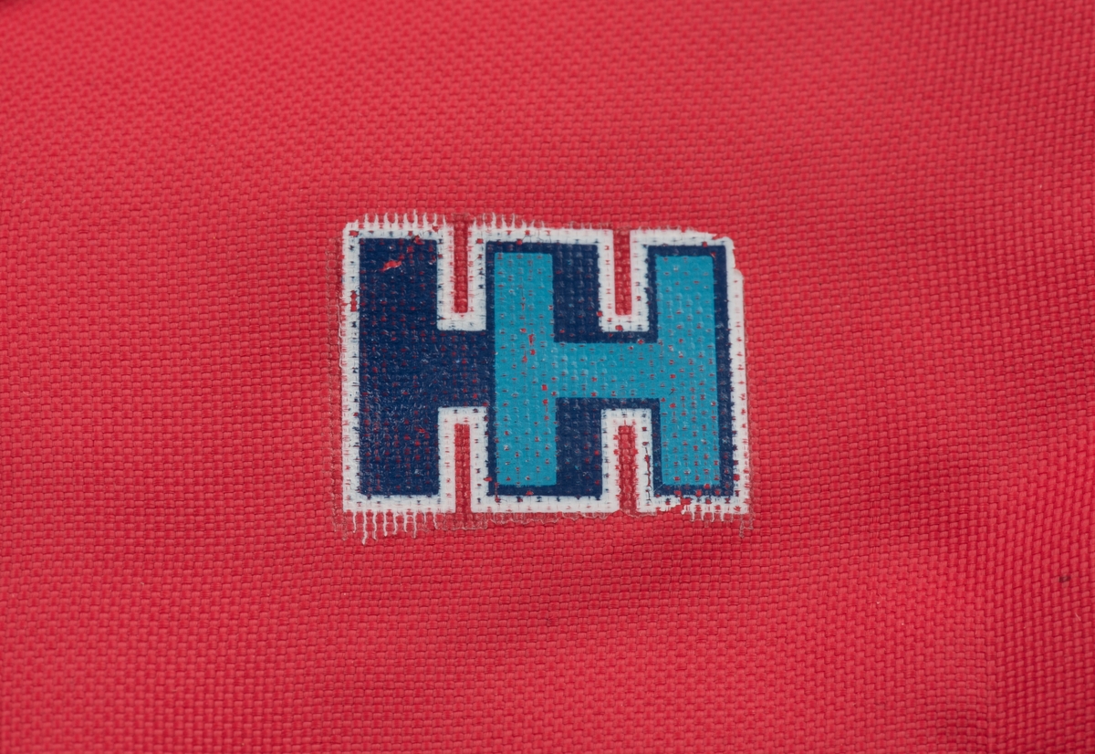 Röda tumvantar av polyester för barn av märket Helly Hansen.
Yttertyg av röd polyester och inuti ett foder av vitt fleecetyg.
På vänster vantes utsida finns Helly Hansens märke "HH" i två blåa nyanser med vit kant.
Vantarna är välanvända och jämnt slitna. De etiketter som finns inuti vantarna är blekta och svårlästa.