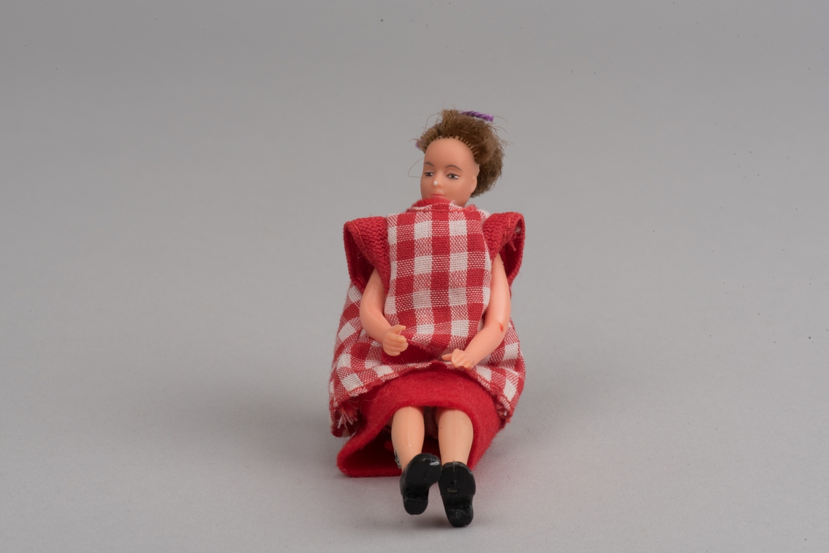 Kvinnlig docka för dockskåp, tillverkad av Lundby leksaksfabrik, i plast.
Dockan är klädd i en röd klänning och ett rödvitrutigt förkläde. Klänningen hålls ihop med en gummisnodd. Håret är ihophållet av en lila tråd i tofs. På fötterna är det målat svarta skor.
Dockan är böjlig. På det ena benet har plasten gått sönder och metallkonstruktionen under är synlig.