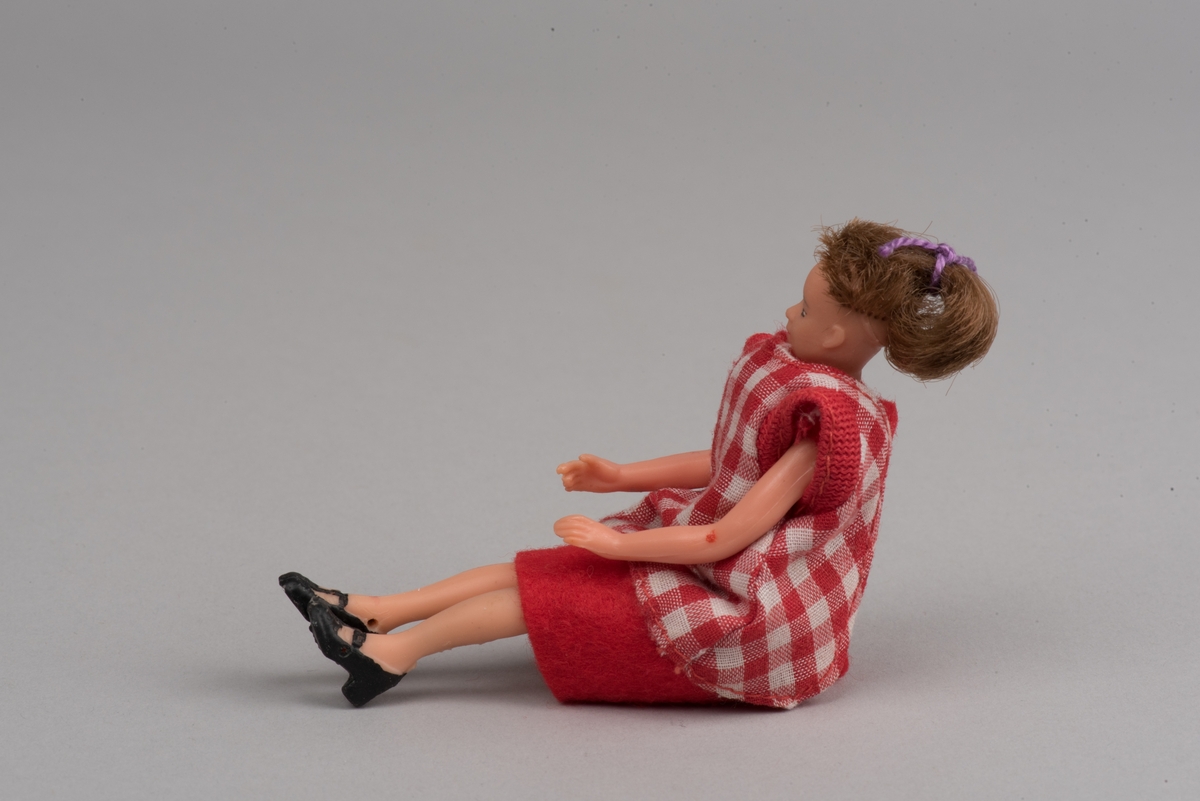 Kvinnlig docka för dockskåp, tillverkad av Lundby leksaksfabrik, i plast.
Dockan är klädd i en röd klänning och ett rödvitrutigt förkläde. Klänningen hålls ihop med en gummisnodd. Håret är ihophållet av en lila tråd i tofs. På fötterna är det målat svarta skor.
Dockan är böjlig. På det ena benet har plasten gått sönder och metallkonstruktionen under är synlig.