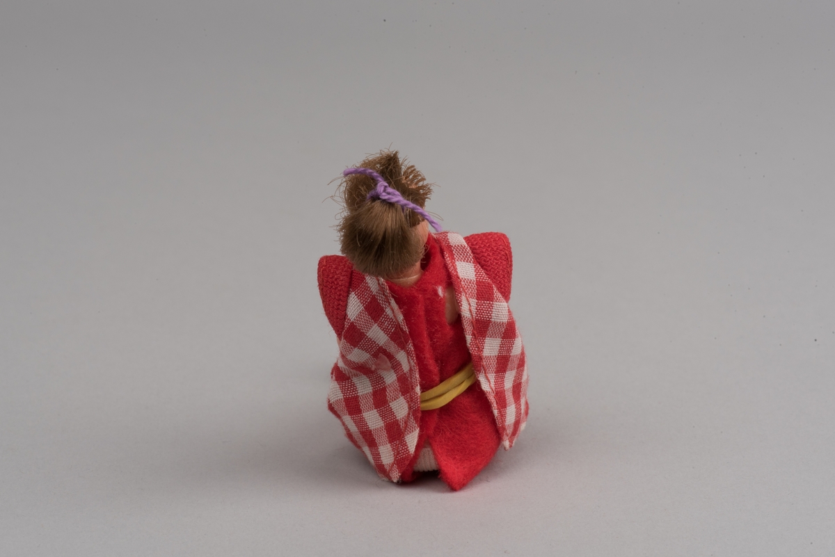 Kvinnlig docka för dockskåp, tillverkad av Lundby leksaksfabrik, i plast.
Dockan är klädd i en röd klänning och ett rödvitrutigt förkläde. Klänningen hålls ihop med en gummisnodd. Håret är ihophållet av en lila tråd i tofs. På fötterna är det målat svarta skor.
Dockan är böjlig. På det ena benet har plasten gått sönder och metallkonstruktionen under är synlig.