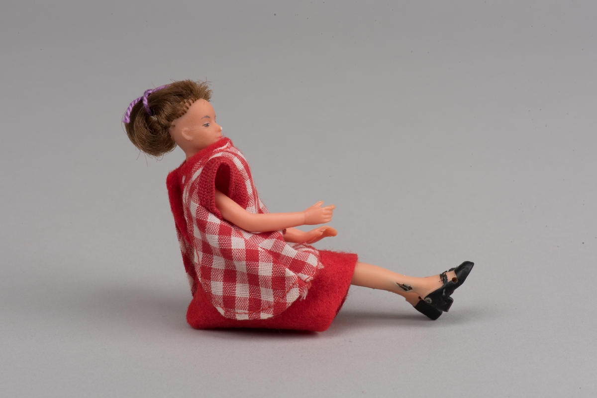 Kvinnlig docka för dockskåp, tillverkad av Lundby leksaksfabrik, i plast.
Dockan är klädd i en röd klänning och ett rödvitrutigt förkläde. Klänningen hålls ihop med en gummisnodd. Håret är ihophållet av en lila tråd i tofs. På fötterna är det målat svarta skor.
Dockan är böjlig. På det ena benet har plasten gått sönder och metallkonstruktionen under är synlig.