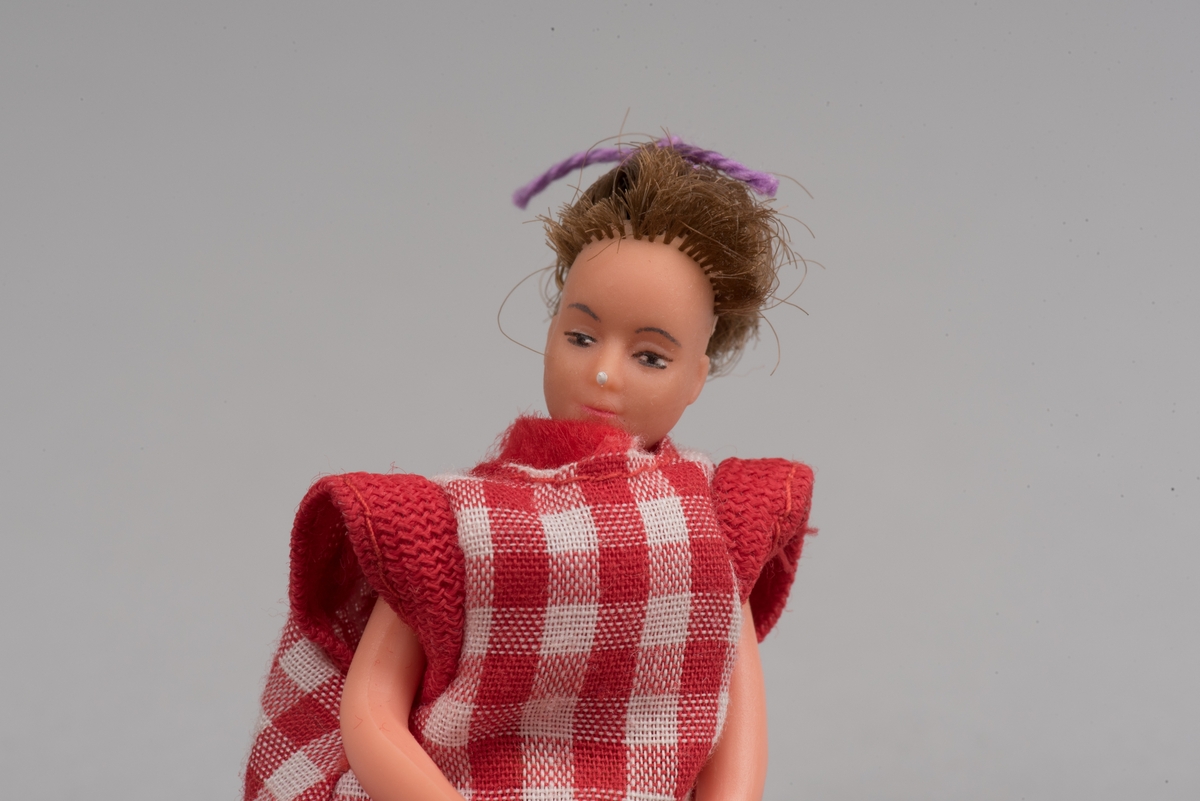 Kvinnlig docka för dockskåp, tillverkad av Lundby leksaksfabrik, i plast.
Dockan är klädd i en röd klänning och ett rödvitrutigt förkläde. Klänningen hålls ihop med en gummisnodd. Håret är ihophållet av en lila tråd i tofs. På fötterna är det målat svarta skor.
Dockan är böjlig. På det ena benet har plasten gått sönder och metallkonstruktionen under är synlig.