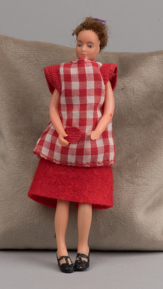 Kvinnlig docka för dockskåp, tillverkad av Lundby leksaksfabrik, i plast.
Dockan är klädd i en röd klänning och ett rödvitrutigt förkläde. Klänningen hålls ihop med en gummisnodd. Håret är ihophållet av en lila tråd i tofs. På fötterna är det målat svarta skor.
Dockan är böjlig. På det ena benet har plasten gått sönder och metallkonstruktionen under är synlig.