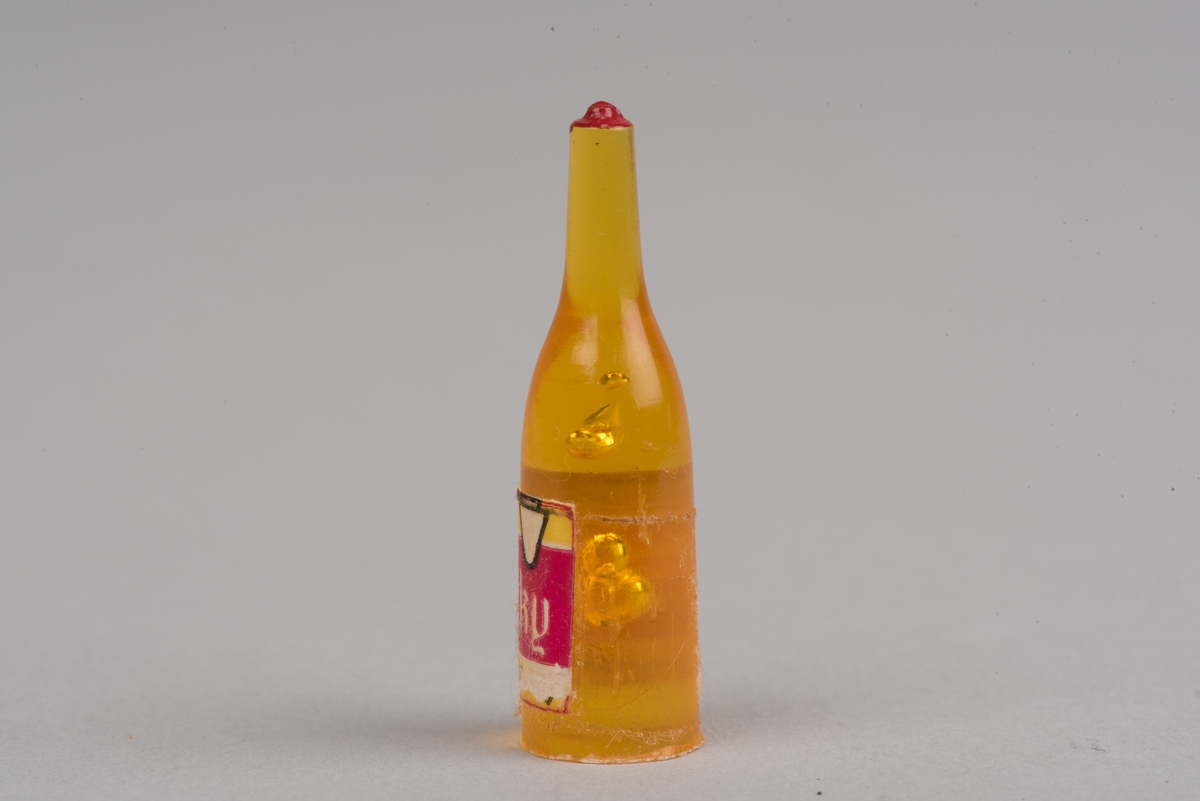 Dockskåpsinredning i form av en whiskyflaska i plast med pappersetikett.
Flaskan är gul med en röd kork. På etiketten framgår att det ska vara en whiskyflaska.