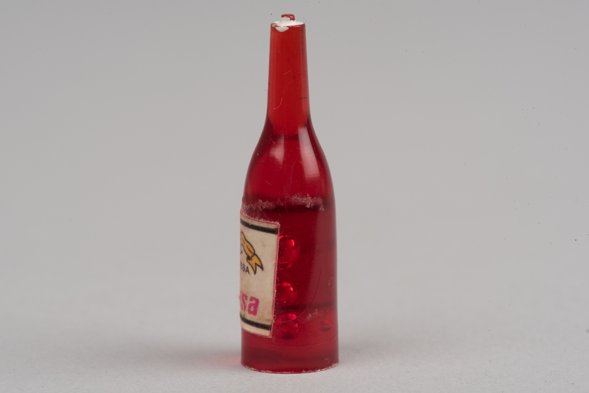 Dockskåpsinredning i form av en vinflaska av plast med etikett.
Flaskan är röd med en vit kork. På etiketten framkommer det att det ska vara en rödvinflaska från Gandesa i Spanien. Etiketten har text och en vindruvsklase.