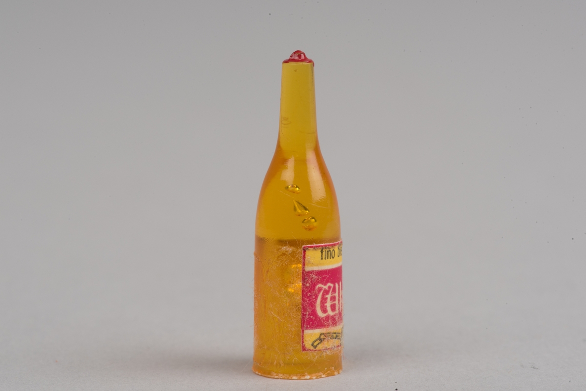 Dockskåpsinredning i form av en whiskyflaska i plast med pappersetikett.
Flaskan är gul med en röd kork. På etiketten framgår att det ska vara en whiskyflaska.