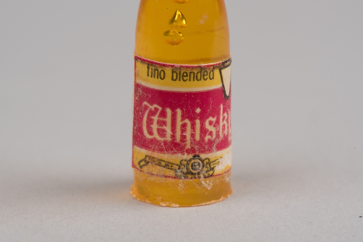 Dockskåpsinredning i form av en whiskyflaska i plast med pappersetikett.
Flaskan är gul med en röd kork. På etiketten framgår att det ska vara en whiskyflaska.