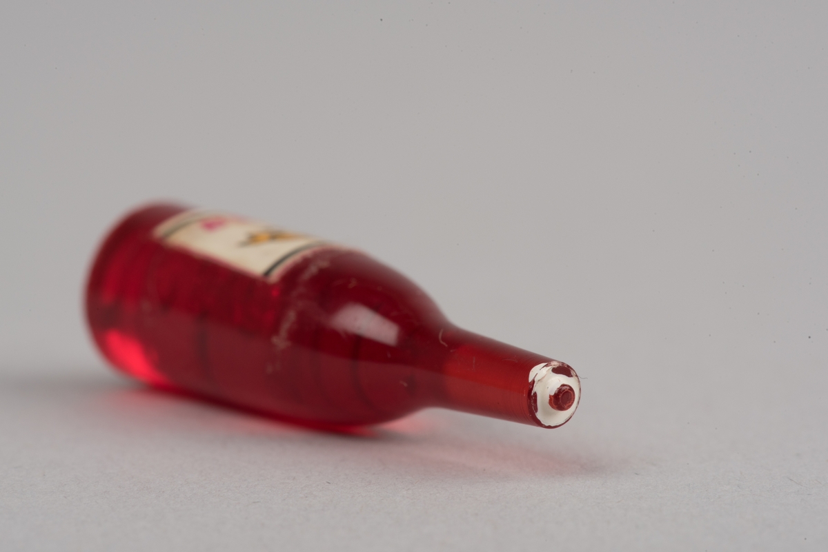 Dockskåpsinredning i form av en vinflaska av plast med etikett.
Flaskan är röd med en vit kork. På etiketten framkommer det att det ska vara en rödvinflaska från Gandesa i Spanien. Etiketten har text och en vindruvsklase.