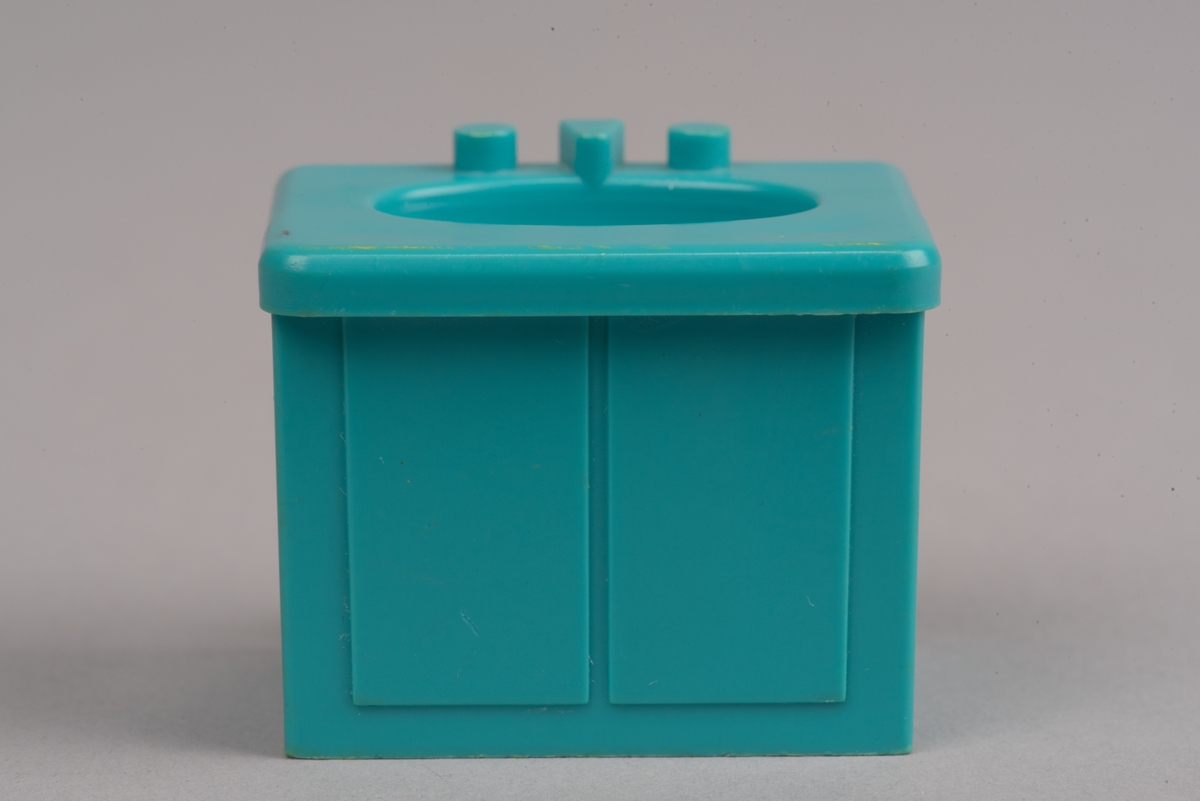 Dockskåpinredning i form av ett turkost tvättställ av plast.
Tvättstället är rektangulärt och har två dörrar på framsidan i relief. Handfatet sticker ut lite på framsidan och har en oval försänkning. Bakom denna två kranar och en blandare i förenklad form. I botten ett hål för vattenavrinning.